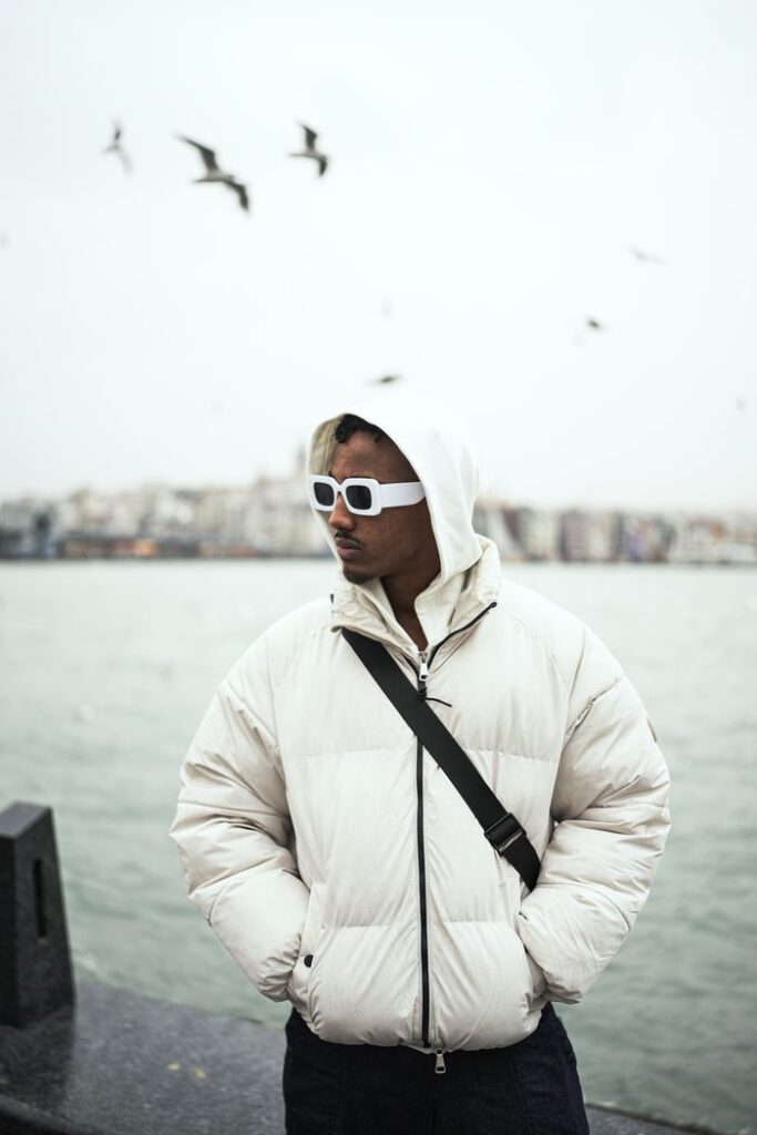 Fashionable man wearing a puffer jacket and sunglasses by a waterfront with birds flying overhead.