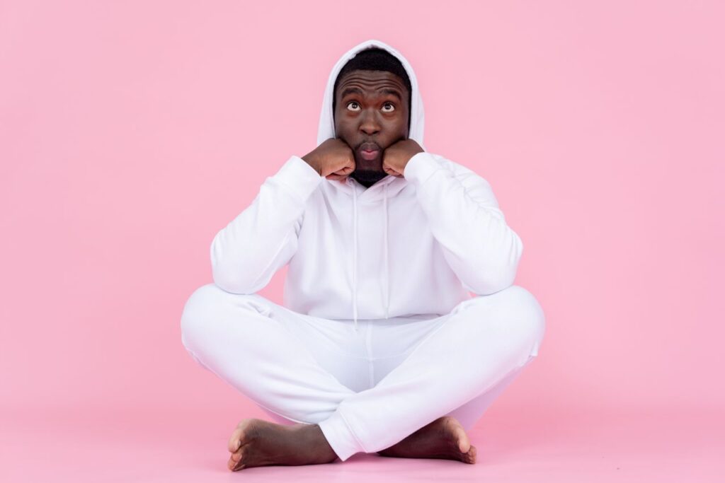 Full body of childish African American male pushing cheeks with fists while pouting lips sitting with crossed legs on floor against pink background